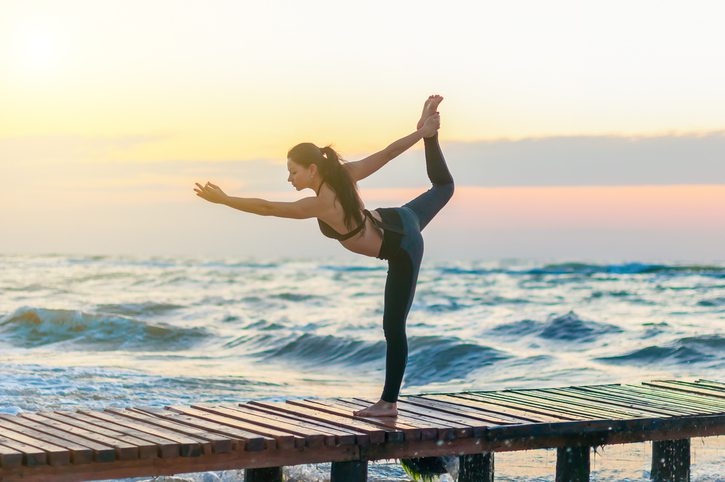 Woman practicing Warrior yoga pose outdoors over sunset sky background.