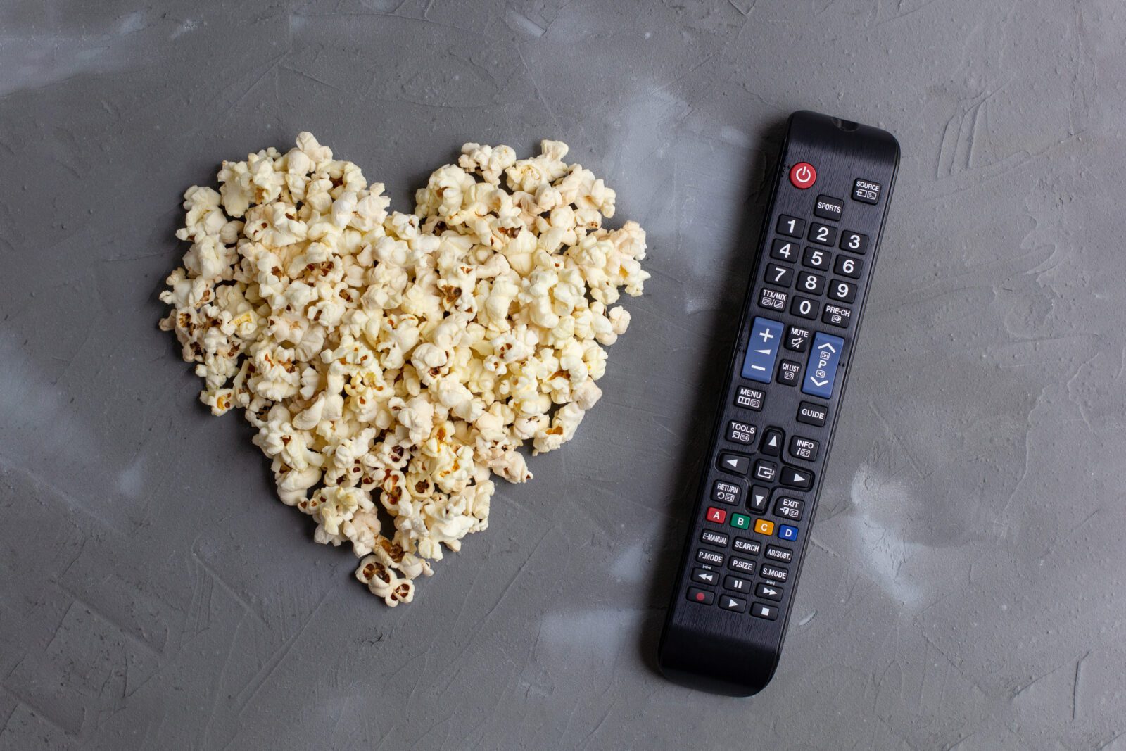 Popcorn in the shape of a heart and TV remote on grey stone background