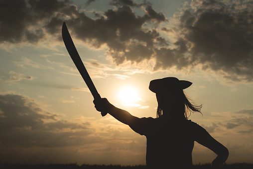 Silhouette of pirate woman in a pirate hat and with a sword in hands over a evening cloudy sky background in sunset rays.